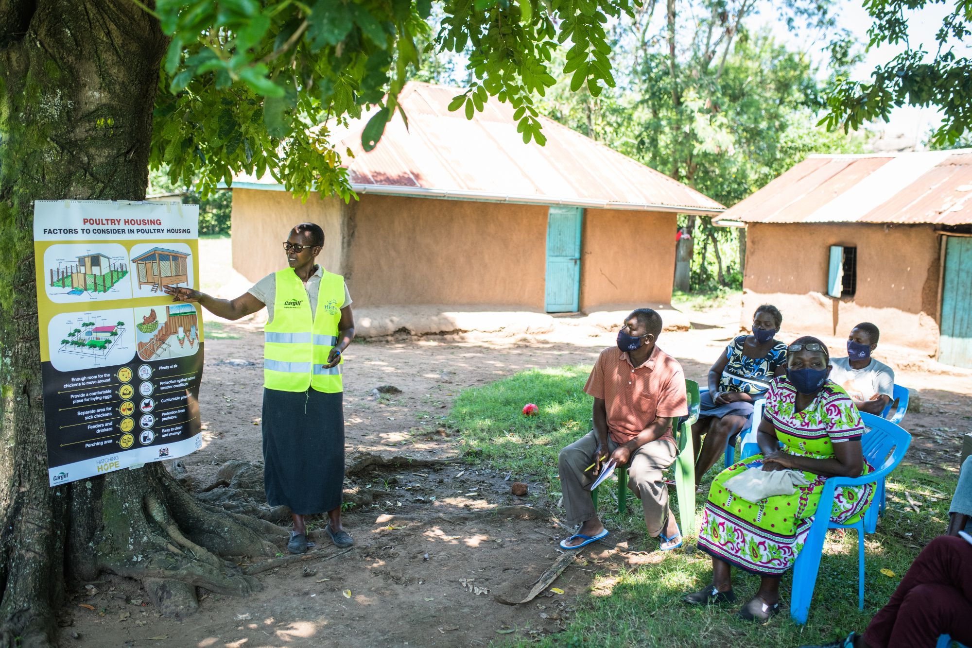 A woman conducting a training with the help of a chart in an open ground with several farmers seated on chairs.