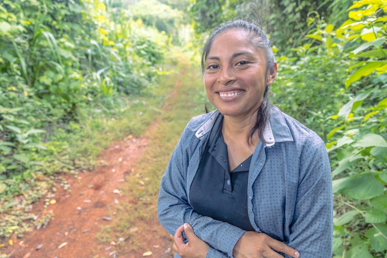 A woman in a dark collared shirt and light gray jacket stands on a dirt path.