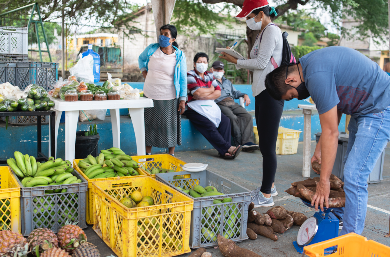 A shot of a local market shows people weighing produce and looking at fruits and vegetables displayed on tables.