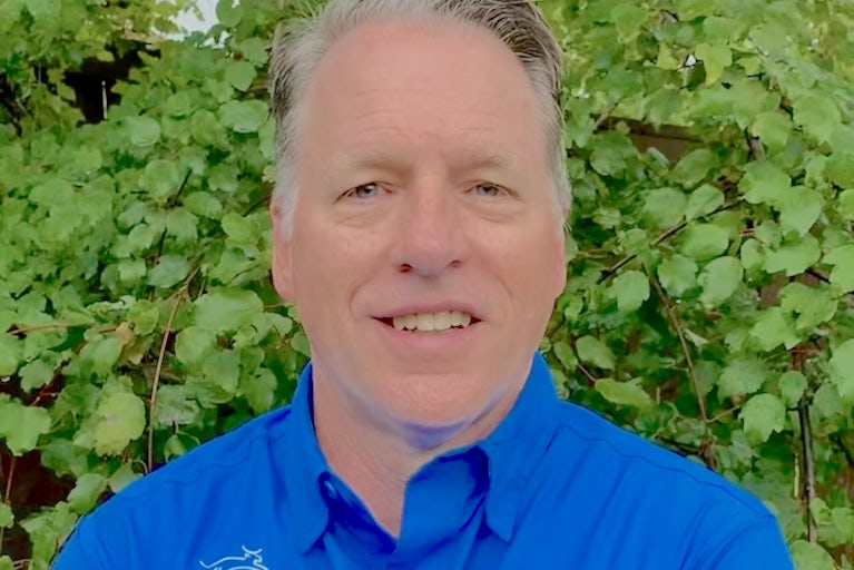 Headshot of Mike Heald wearing a blue Heifer International button-up shirt in front of lush leaves in the background