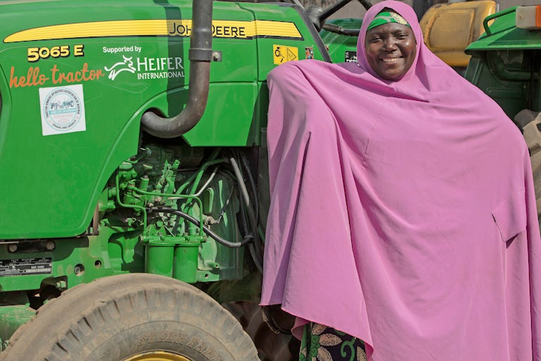 A woman in a pink hijab stands next to a green John Deere tractor.