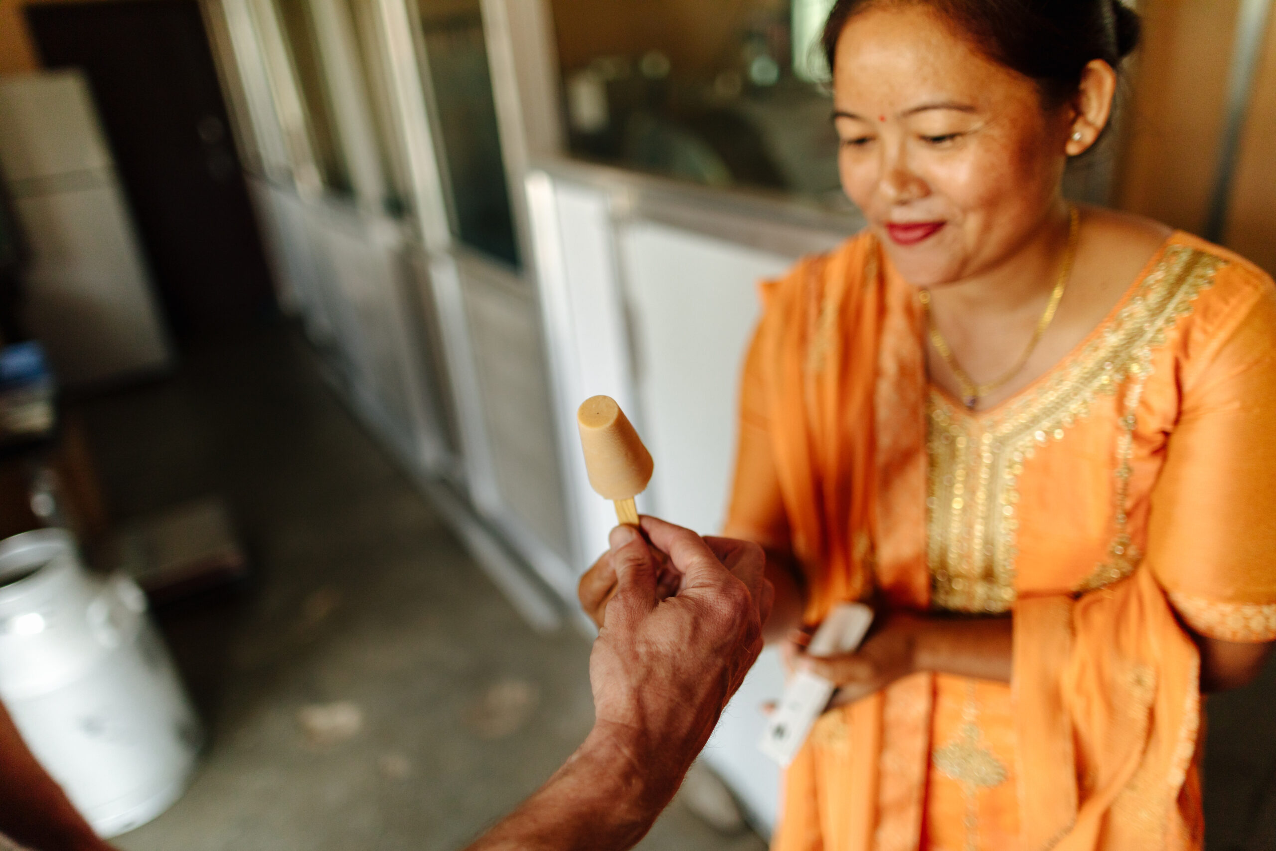 A nepali woman in bright orange kurta suruwal hands an ice cream to a customer off camera