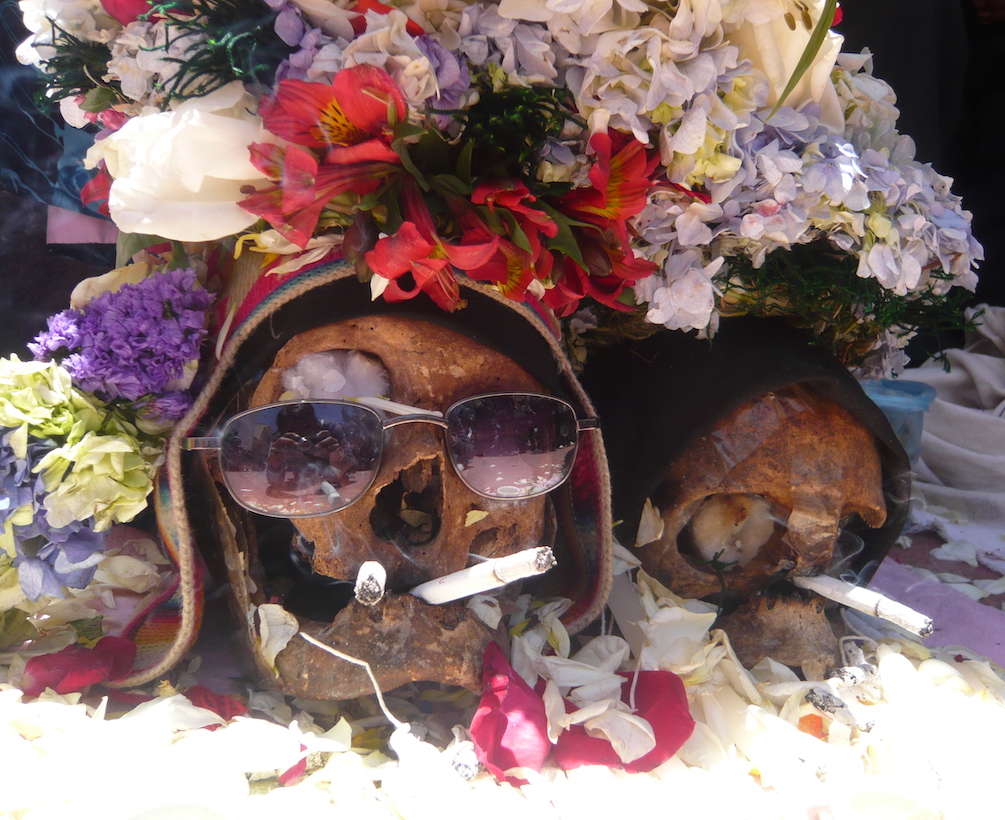 Two skulls adorned in flowers sit on the ground with lit cigarettes in their mouths.