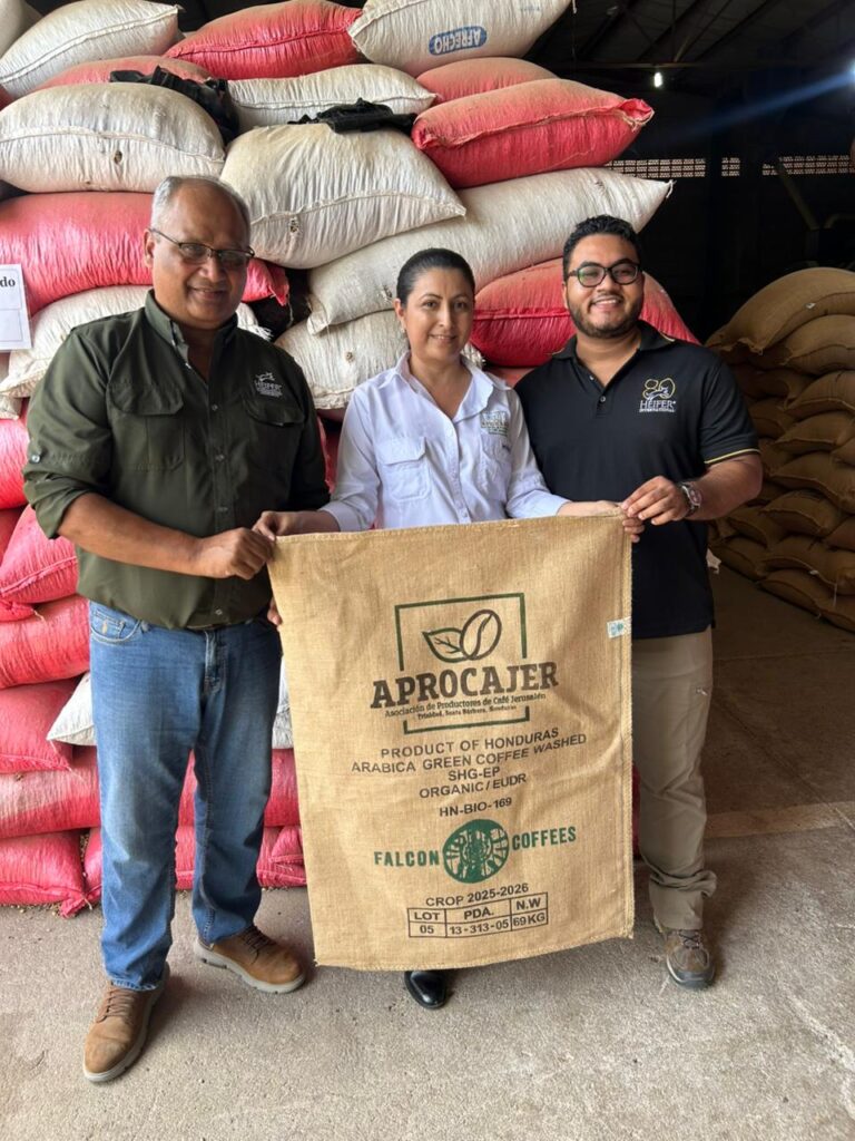 A woman and two men hold an APROCAJER coffee sack in front of stacks of bagged coffee.