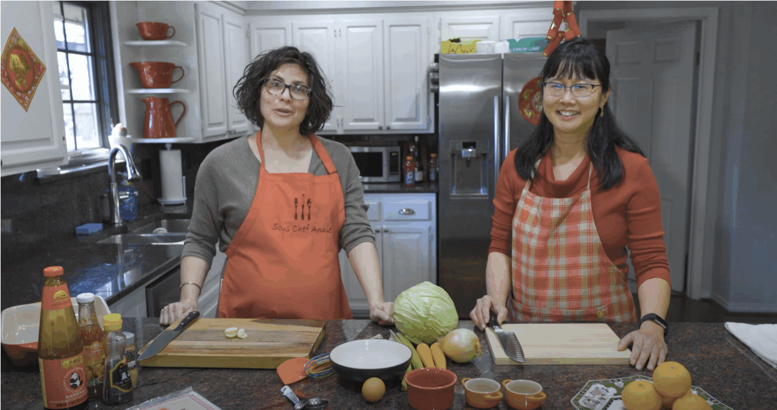 Two women, in red aprons, stand smiling for the camera. They are standing behind a kitchen island.