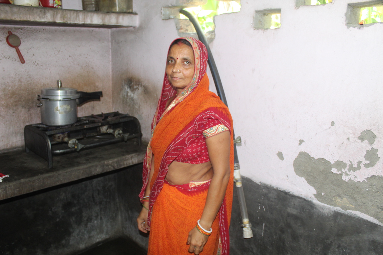 An Indian woman stands in front of her stove, looking at the camera.