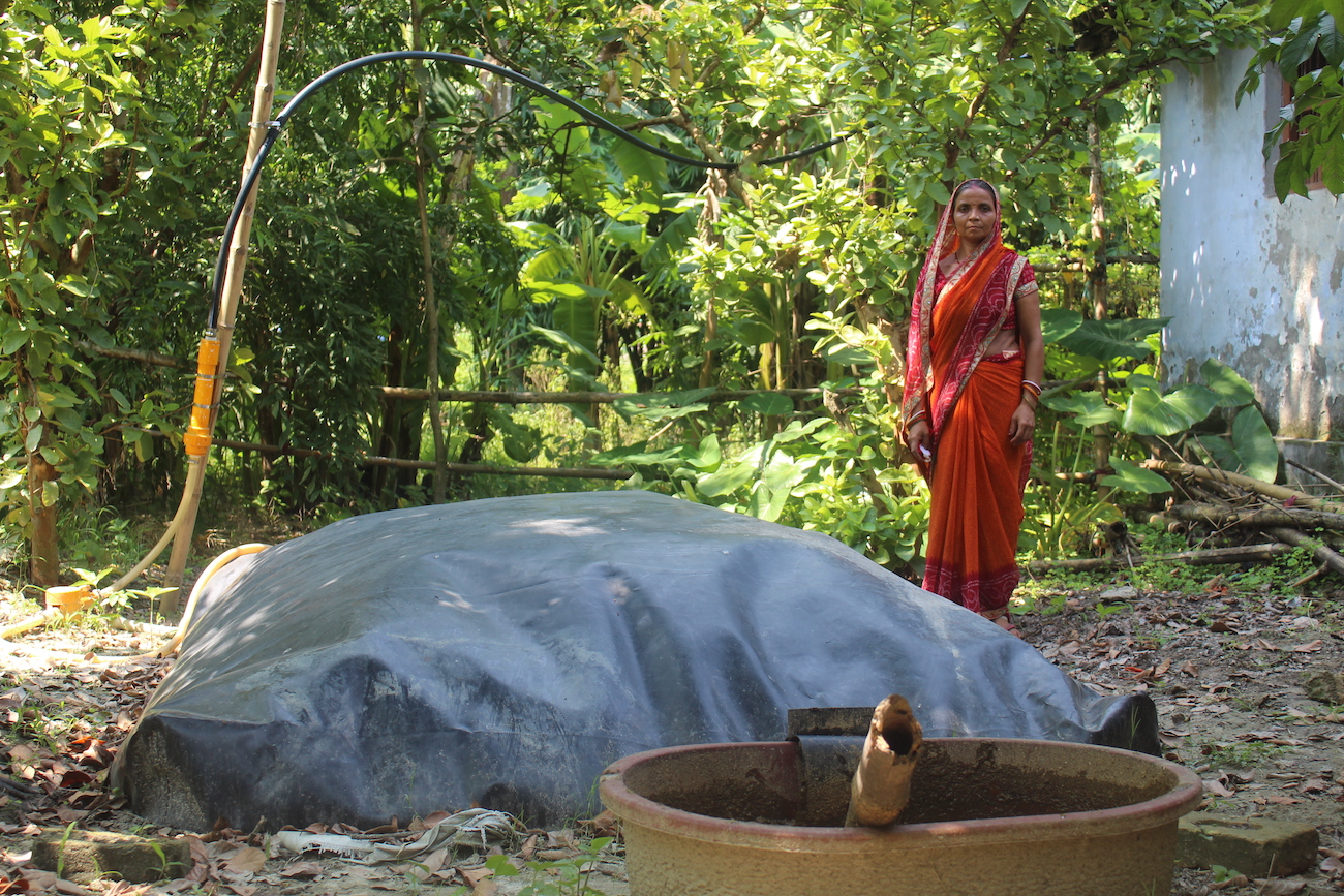 An Indian woman in orange dress stands behind her biodigester, a large grey dome on the ground.