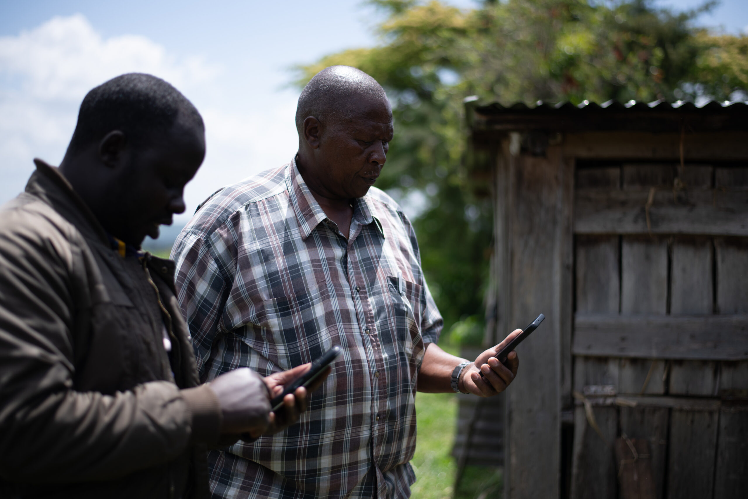 Two men stand outdoors and check information on their mobile phones.