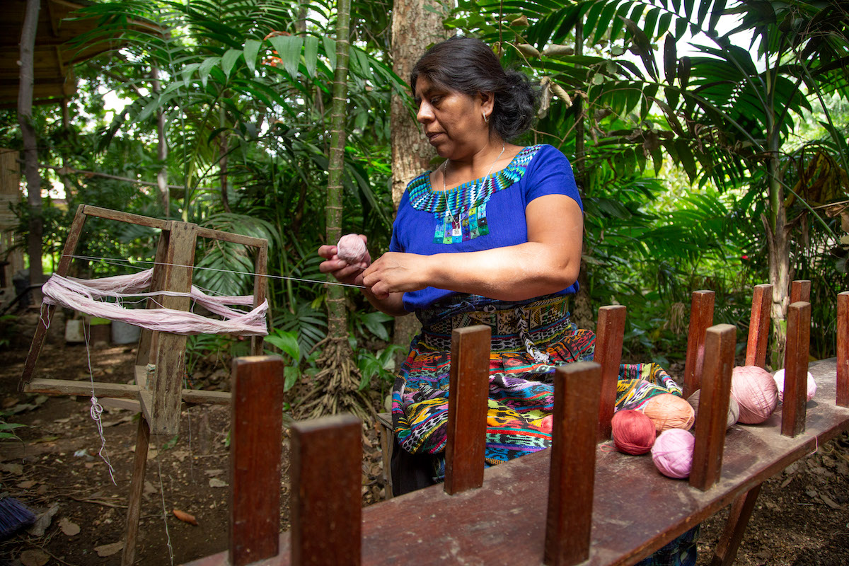 Rosalinda Tay sits outside her studio and winds yarn died with cochineal on her enrollado. In front of her is an urdidora or warping frame.