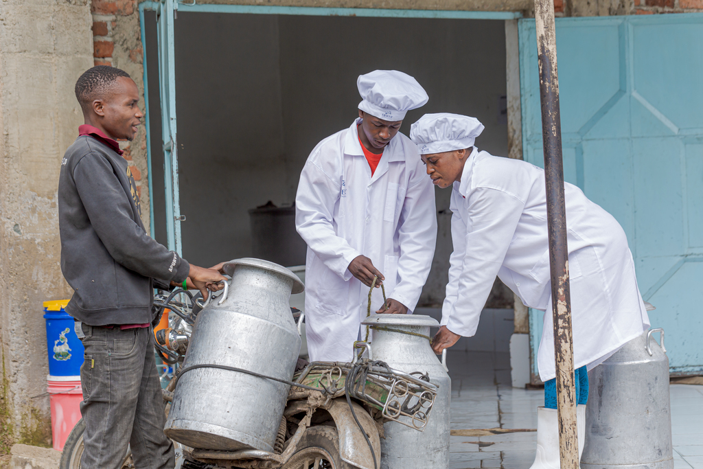 Two workers in lab coats inspect milk containers beside a motorcycle.
