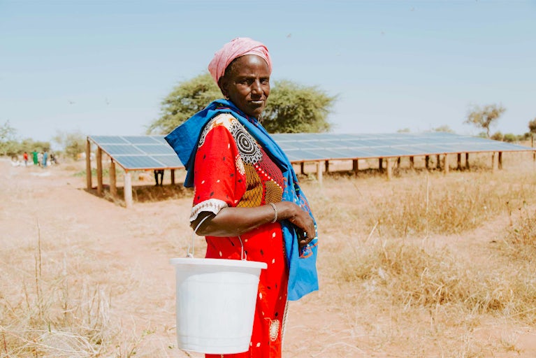 Woman in colorful dress stands by solar panels with water bucket.