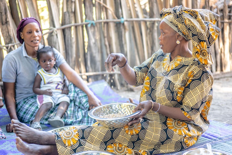 A woman in sunflower-patterned attire educates others on porridge production in a Senegalese village.