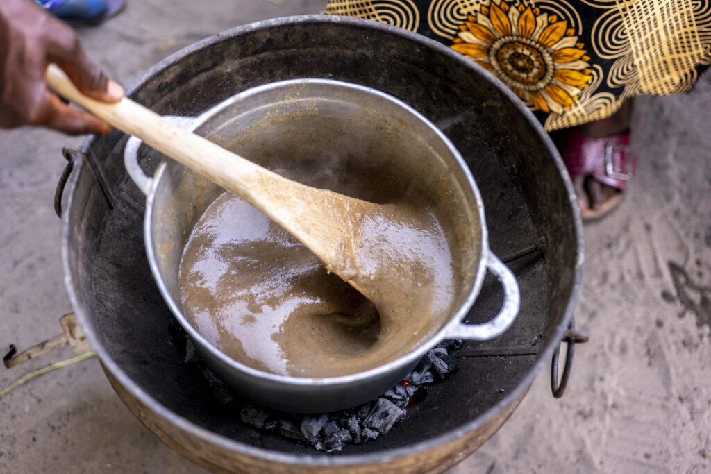 A woman in patterned clothing stirs porridge cooking in a pot over a fire.