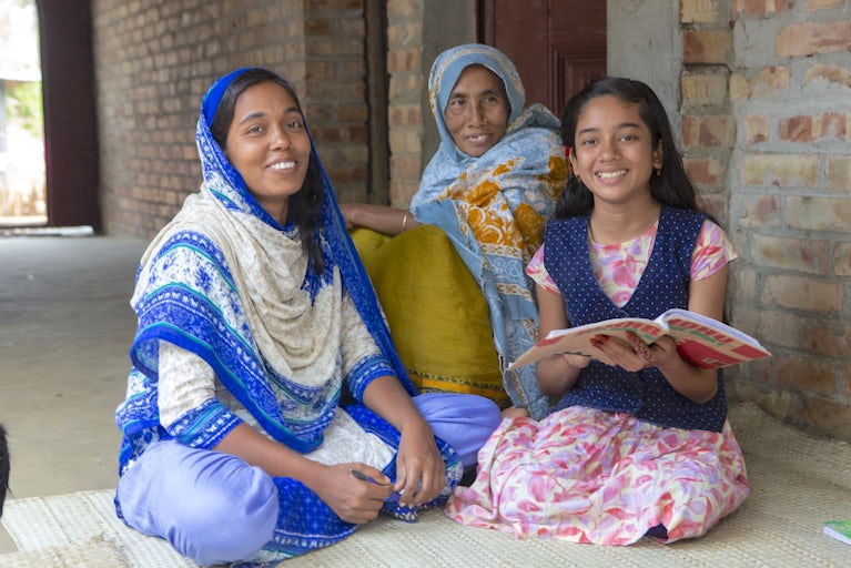 Women sitting on the ground smiling and reading a book.