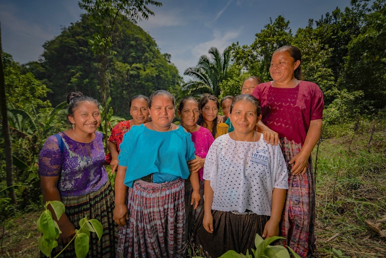 A group of women and girls in traditional attire stand outdoors in a tropical setting.