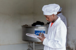 A man in a white coat and hat records notes while standing near dairy equipment.
