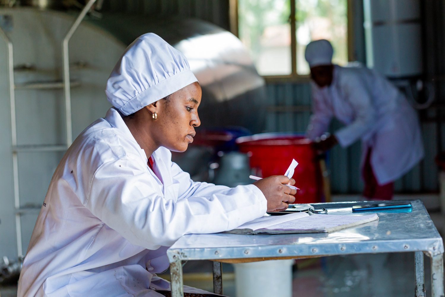 A dairy worker in a white lab coat sits at a table, examining a document, with large metal containers in the background.