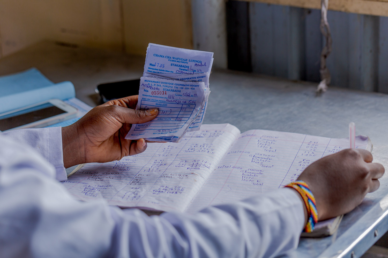 A close-up of a dairy worker's hands holding receipts and writing in a ledger book.