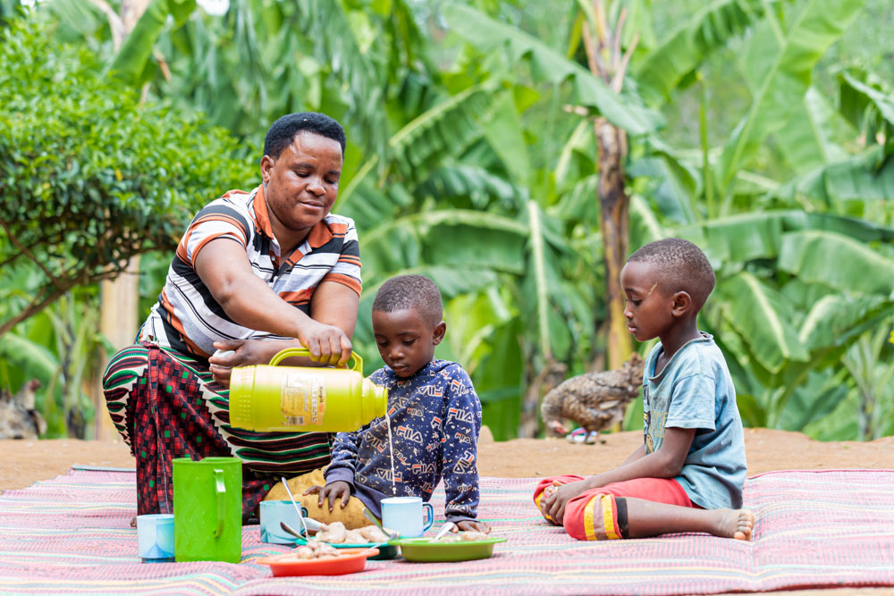 A woman serves milk to her two young children outdoors, surrounded by banana plants.