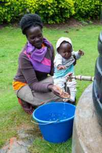 A woman holding her child gathers water from a rainwater collection tank into a blue bucket.