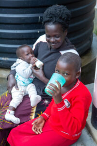 A woman on a step cradles a toddler while another child drinks from a cup.