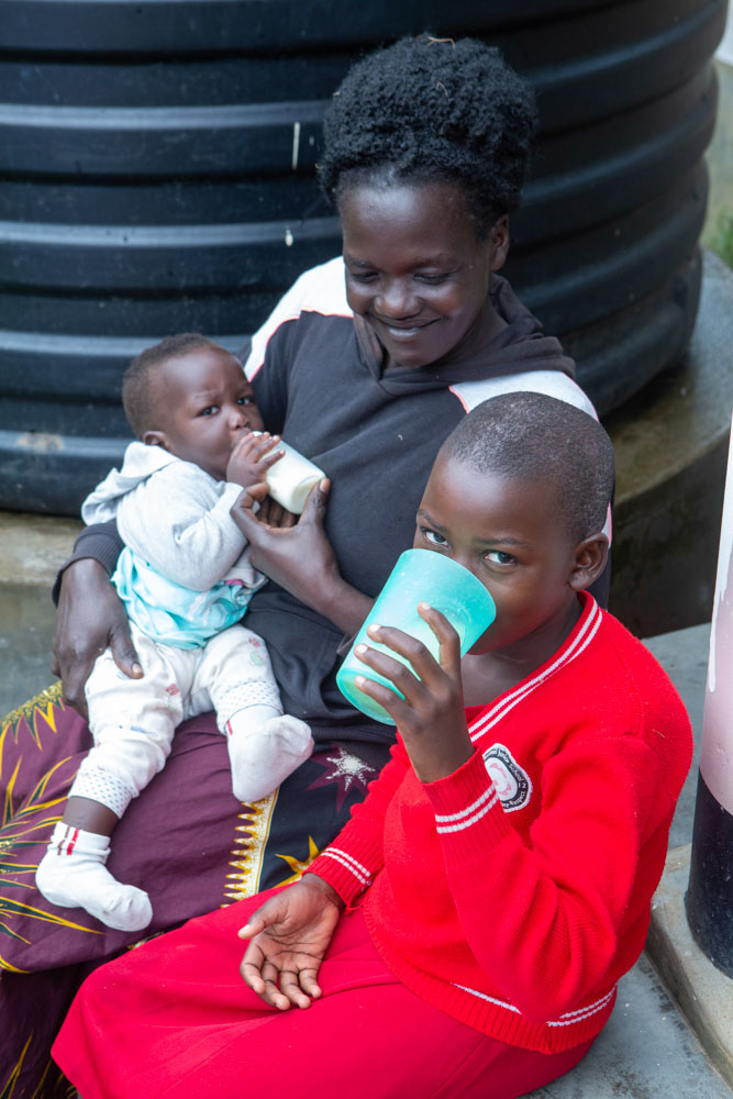 A woman on a step cradles a toddler while another child drinks from a cup.