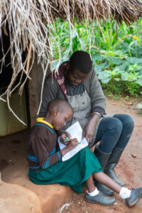 A mother and her daughter sit under a thatched roof, working on school homework together.