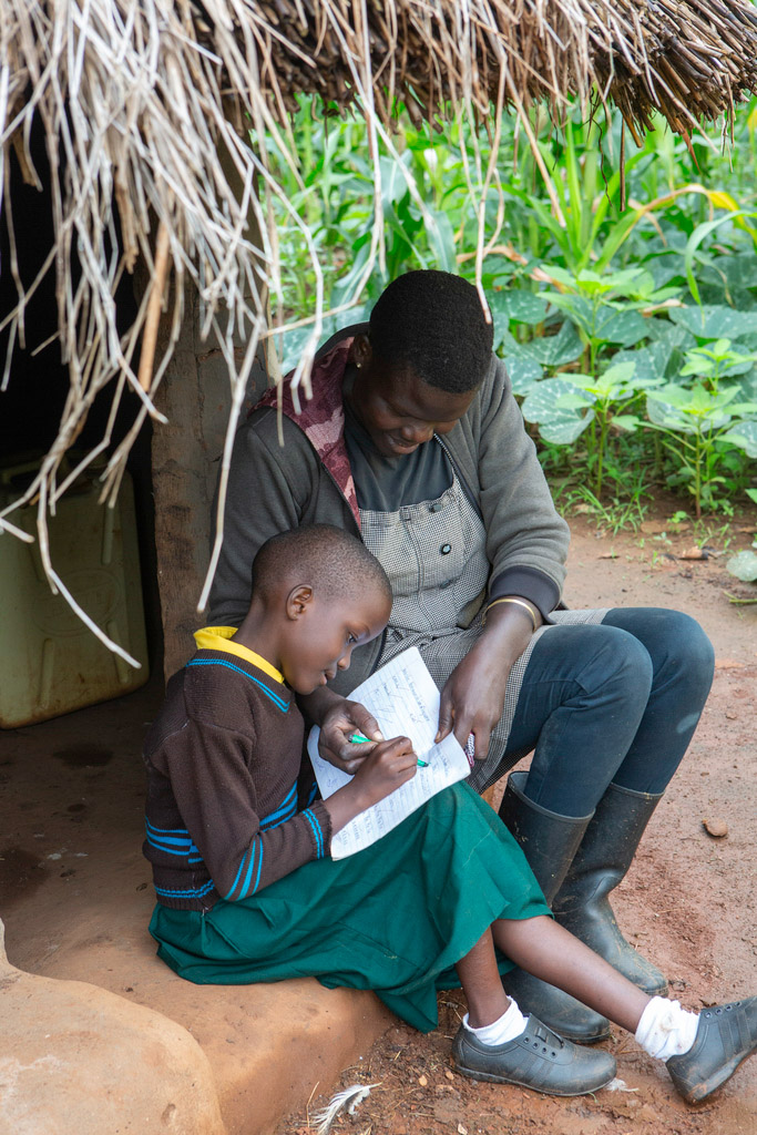 A mother and her daughter sit under a thatched roof, working on school homework together.