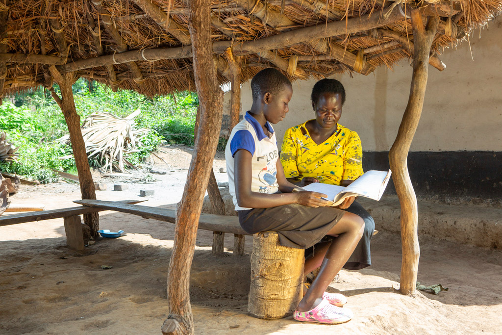 A woman and her daughter smile while reading a book under a thatched canopy.