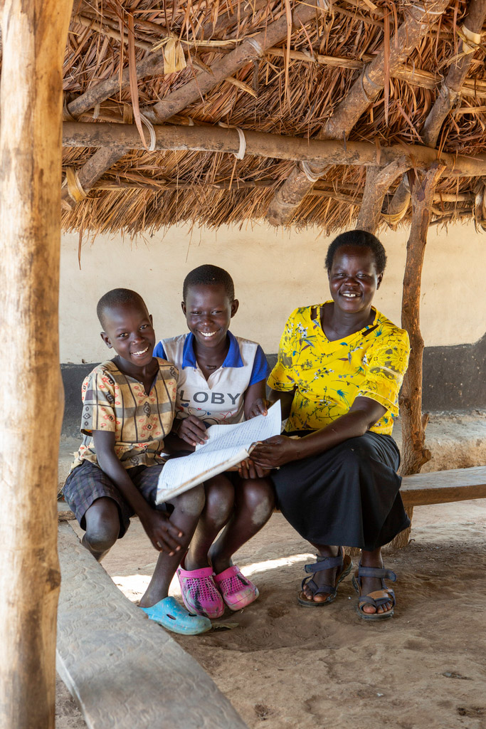 A woman sits with her two children under a thatched canopy, holding a schoolbook.