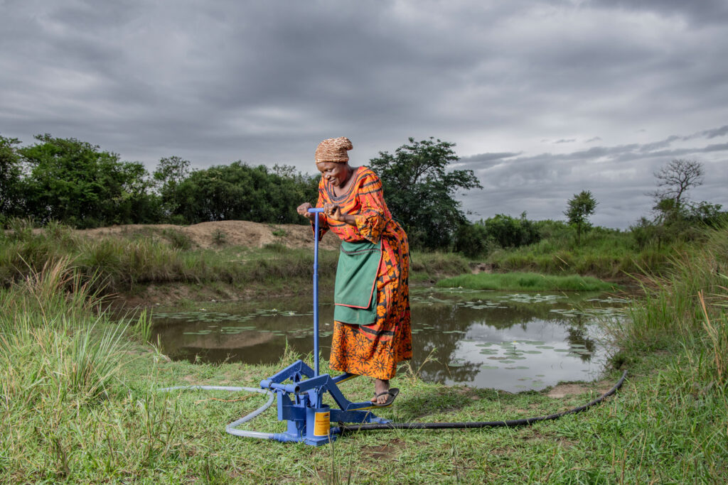 A woman operates a manual water pump beside a dam.