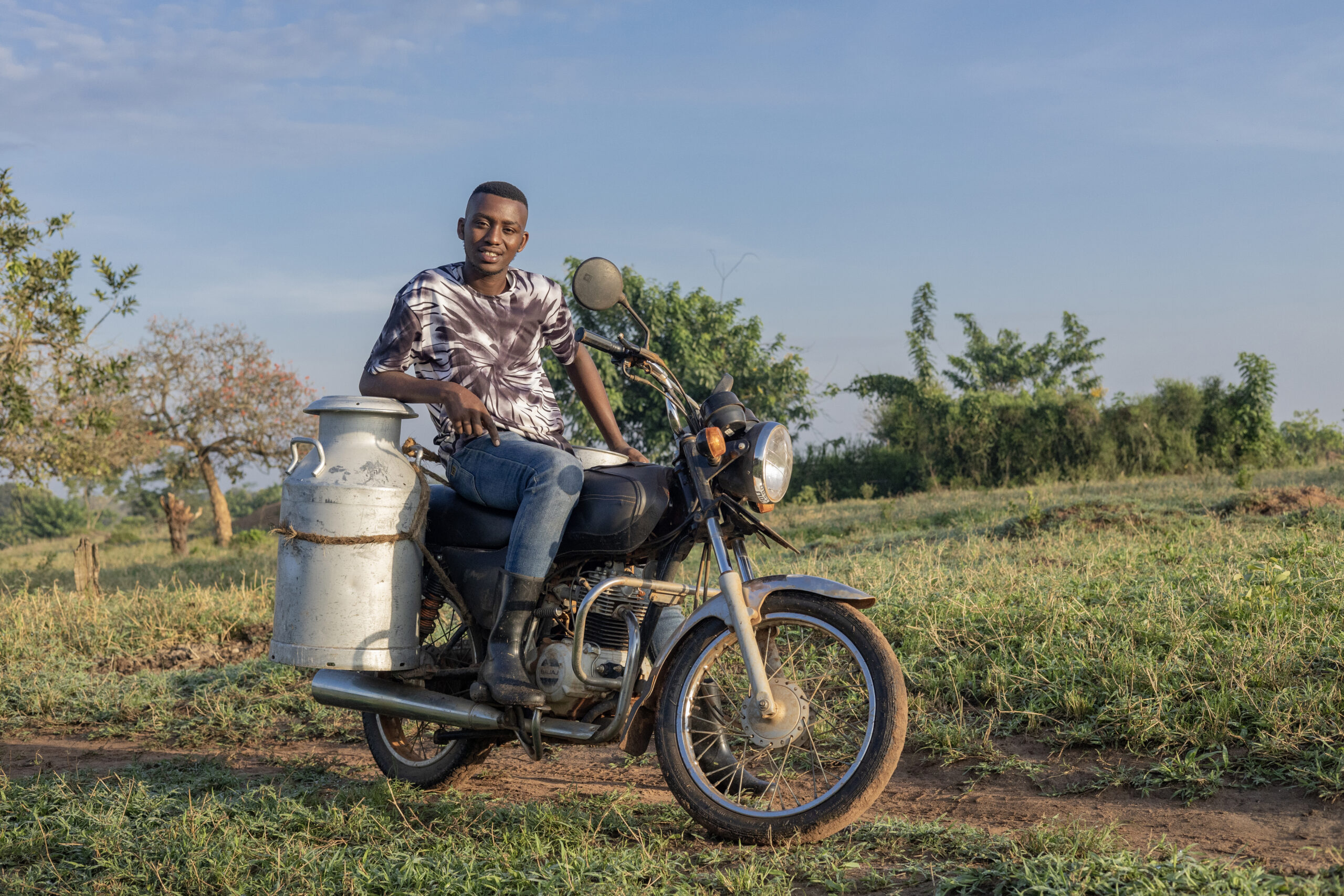 A man sits on a motorcycle with a large milk can strapped to the side.
