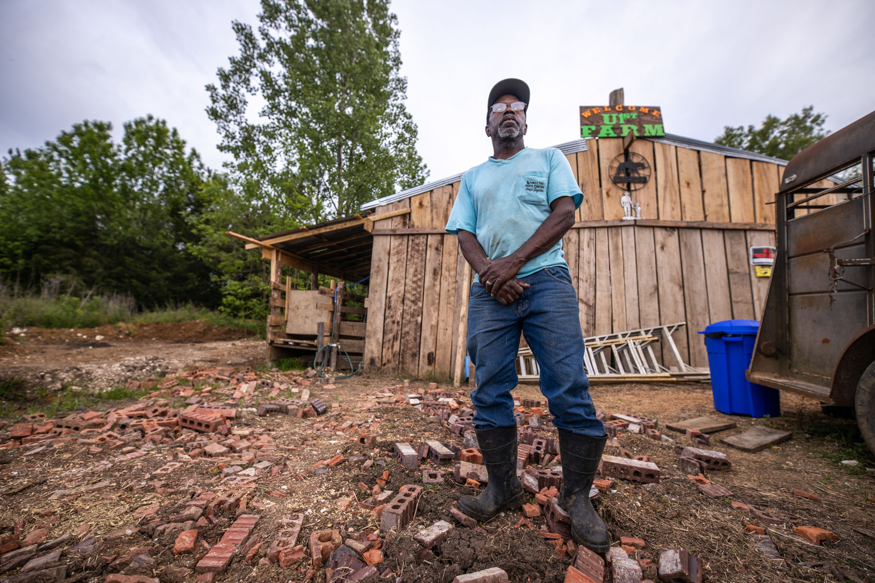 Man in blue shirt standing in front of a wooden farm building. 2 / 2