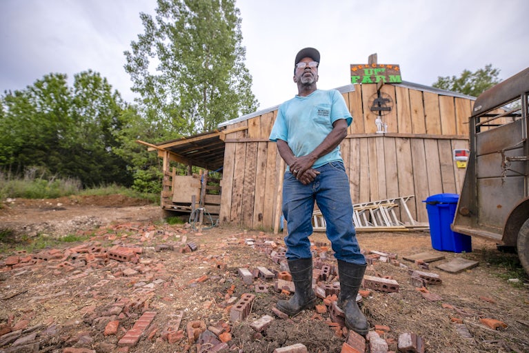 Man in blue shirt standing in front of a wooden farm building. 2 / 2