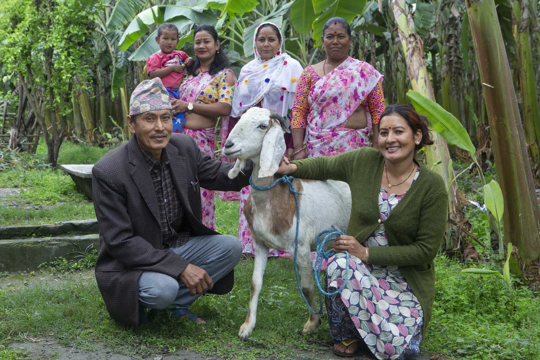 A woman and man crouch next to their goat. Four women stand behind them, smiling.