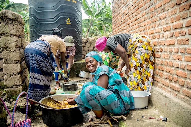 A group of women prepare food together outdoors beside a brick wall and large water tank, with one woman in turquoise clothing smiling brightly at the camera.