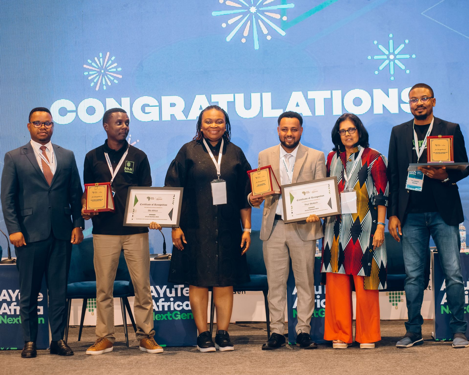 A group of award winners stand on stage holding plaques and certificates at an event.