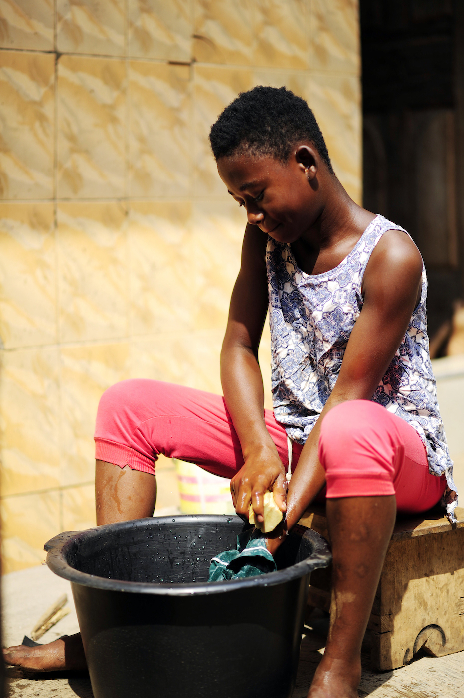 A Ghanese girl sits near a basin, washing her school uniform with the soap her mother made.