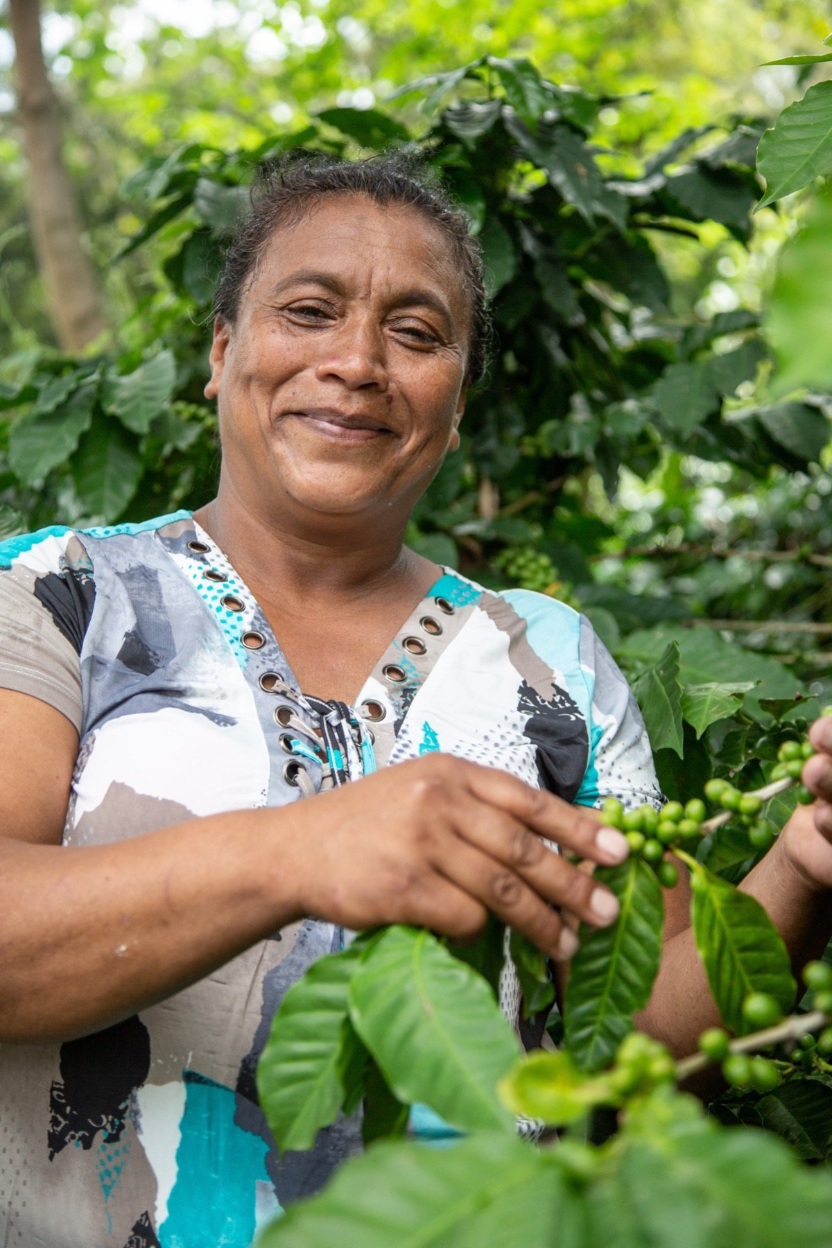 A woman stands next to her coffee plant and smiles at the camera.