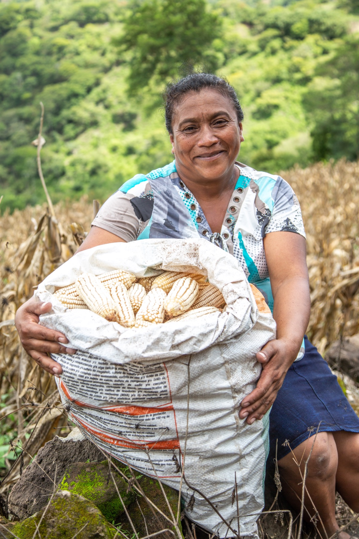 A woman sits in a field, holding a large bag of harvested corn.