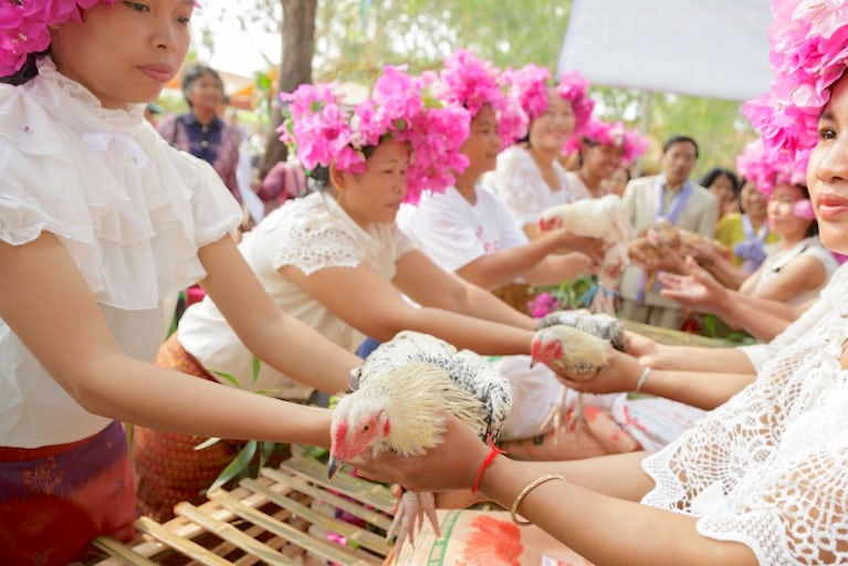 Women in white lace dresses and flower crowns hold chickens during a traditional ceremony.