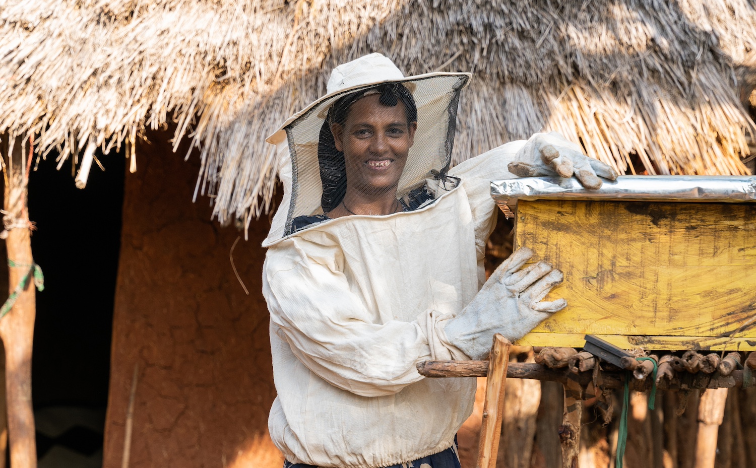A smiling beekeeper in protective gear stands beside a yellow wooden beehive in front of a thatched-roof hut.