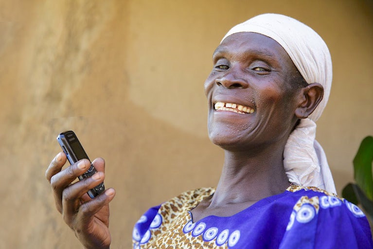Joyful person in white headscarf and blue shirt smiles while holding a mobile phone.