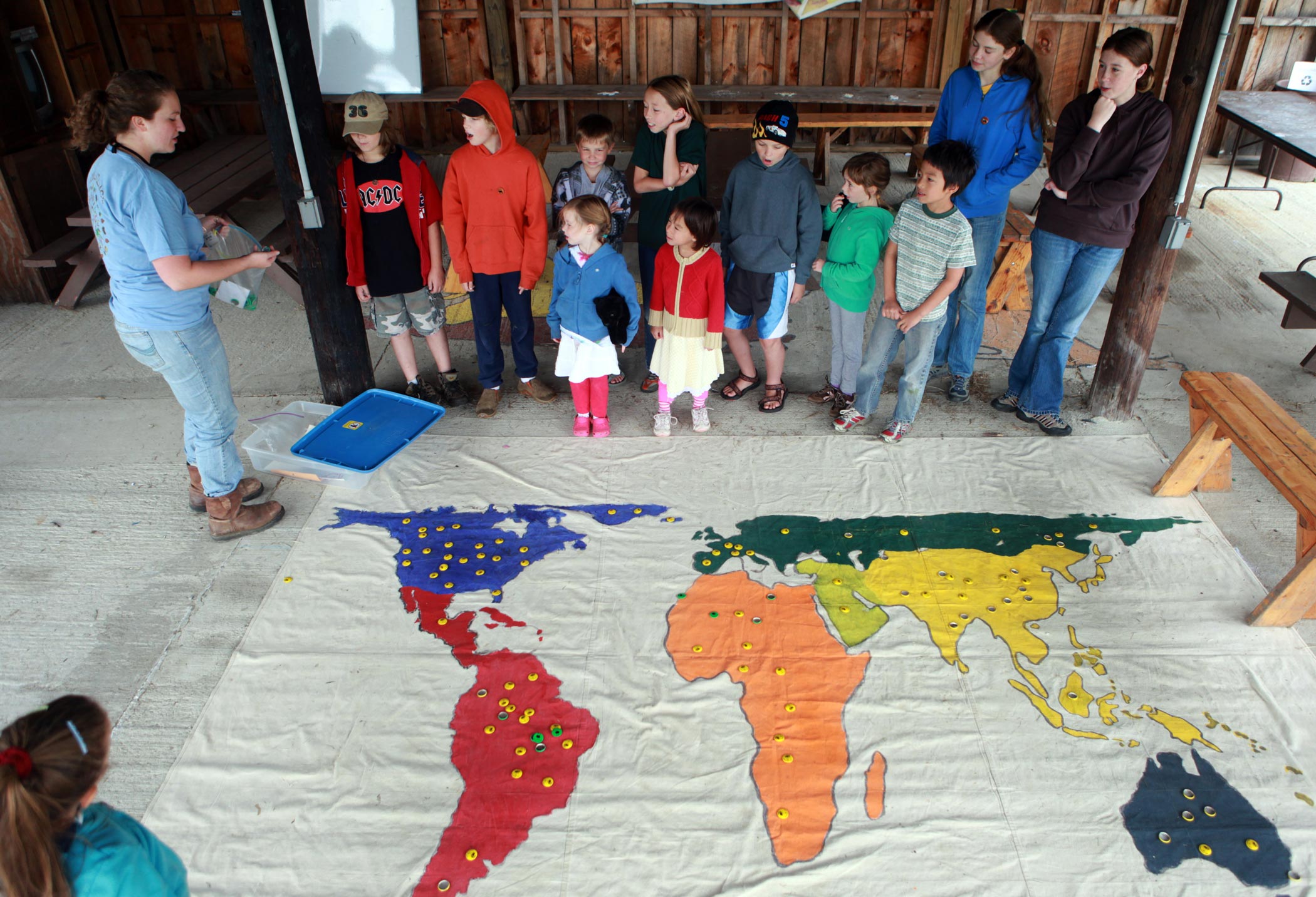Young woman addresses families while standing next to a large, colorful map of Earth on the floor.