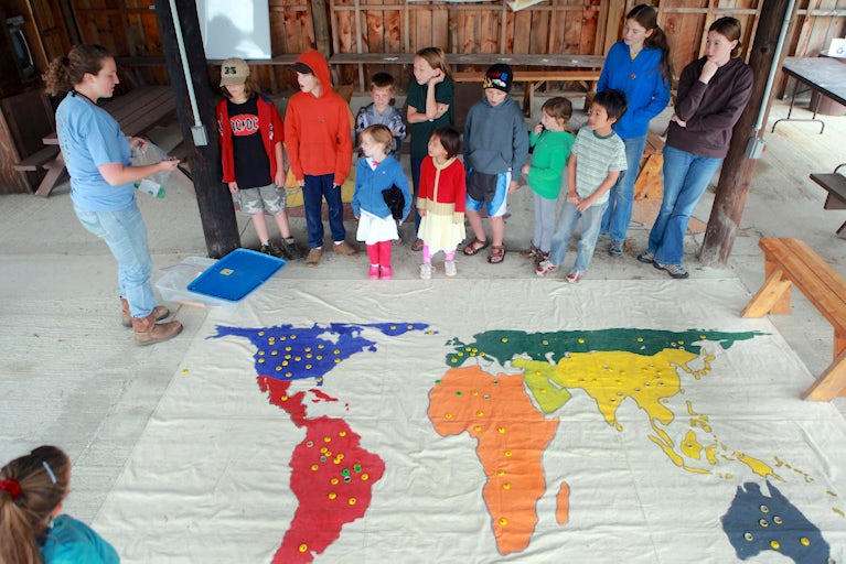 Young woman addresses families while standing next to a large, colorful map of Earth on the floor.