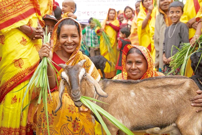 Two women in ceremonial clothing kneeling next to a large goat, surrounded by a crowd of people also dressed in traditional clothing.