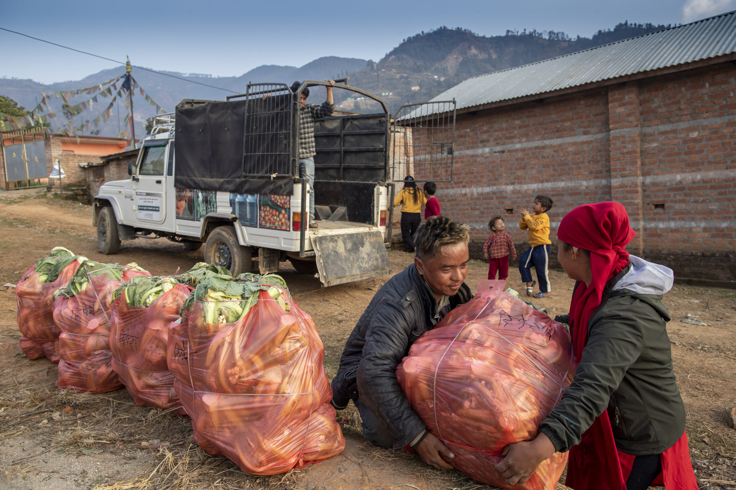 Two people lift a large bag of onions near several similar bags beside a white truck. Children play in the background, with mountains and brick buildings visible under a blue sky.