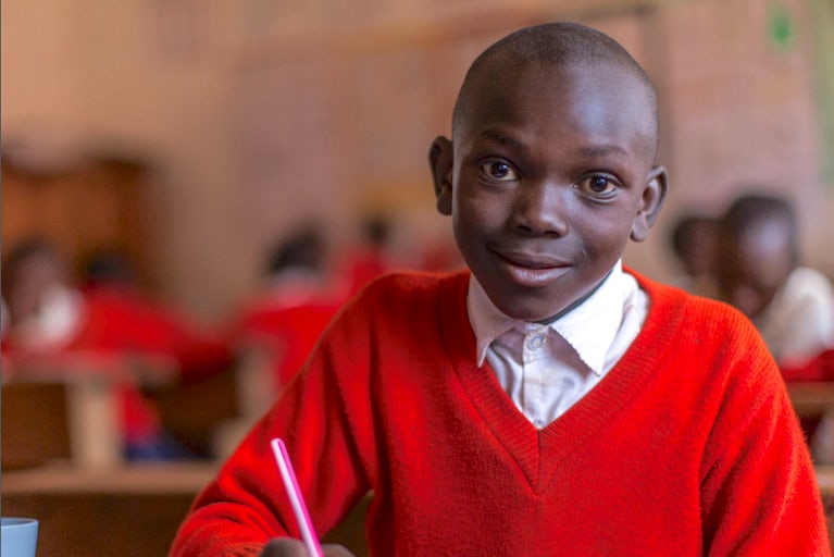 Young boy wearing a red sweater and working on a school assignment in a classroom.