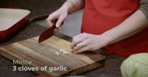 A demonstration shot of mincing garlic - one hand holds a kitchen knife and one holds cloves of garlic.