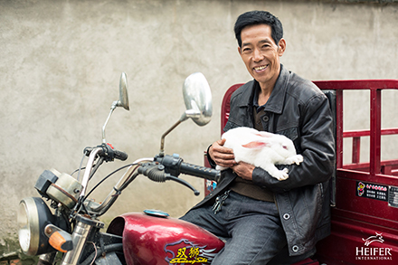 Zhou Xianyun, 49, holds a rabbit as he sits on a motorcycle his family was able to buy thanks to profit from the sale of rabbits. A man on a motorcycle holding a rabbit.